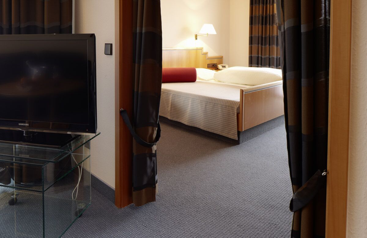 A hotel room with a bed, a TV on a glass stand, a lamp, and a door with curtains.