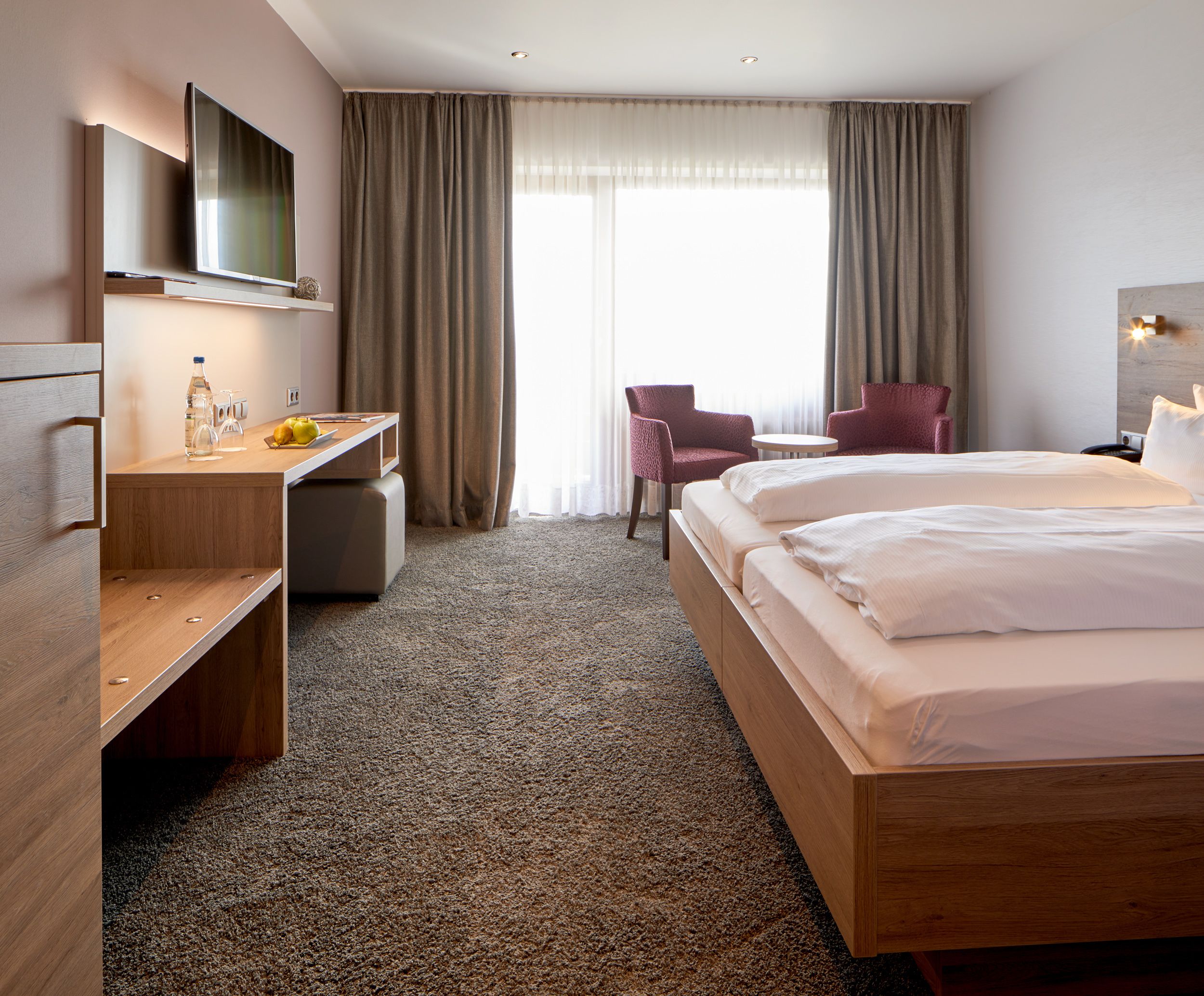 Una habitación de hotel con cama, escritorio, silla, televisión y ventana con cortinas.