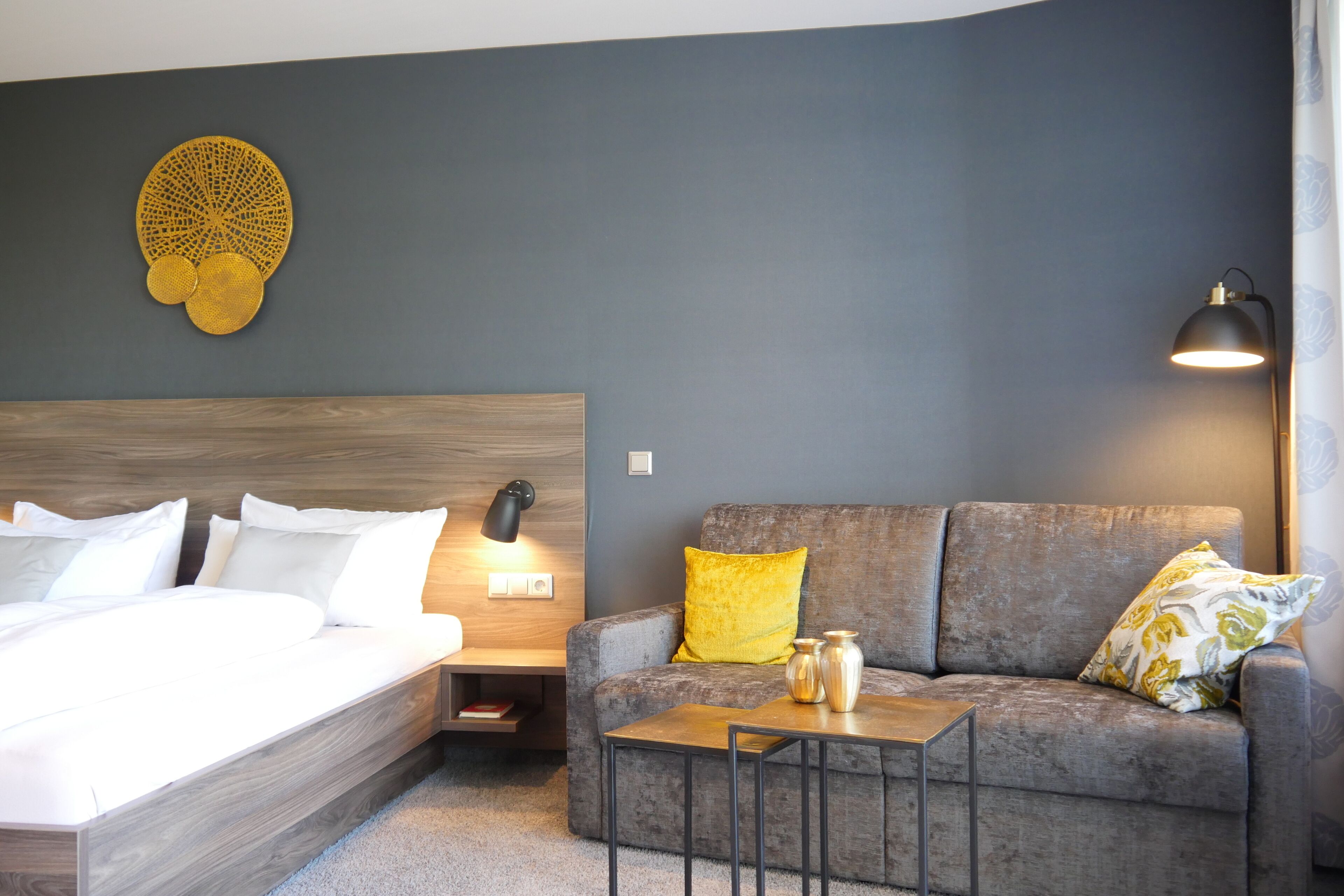A modern hotel room with a wooden bed, a grey sofa, and a small table.