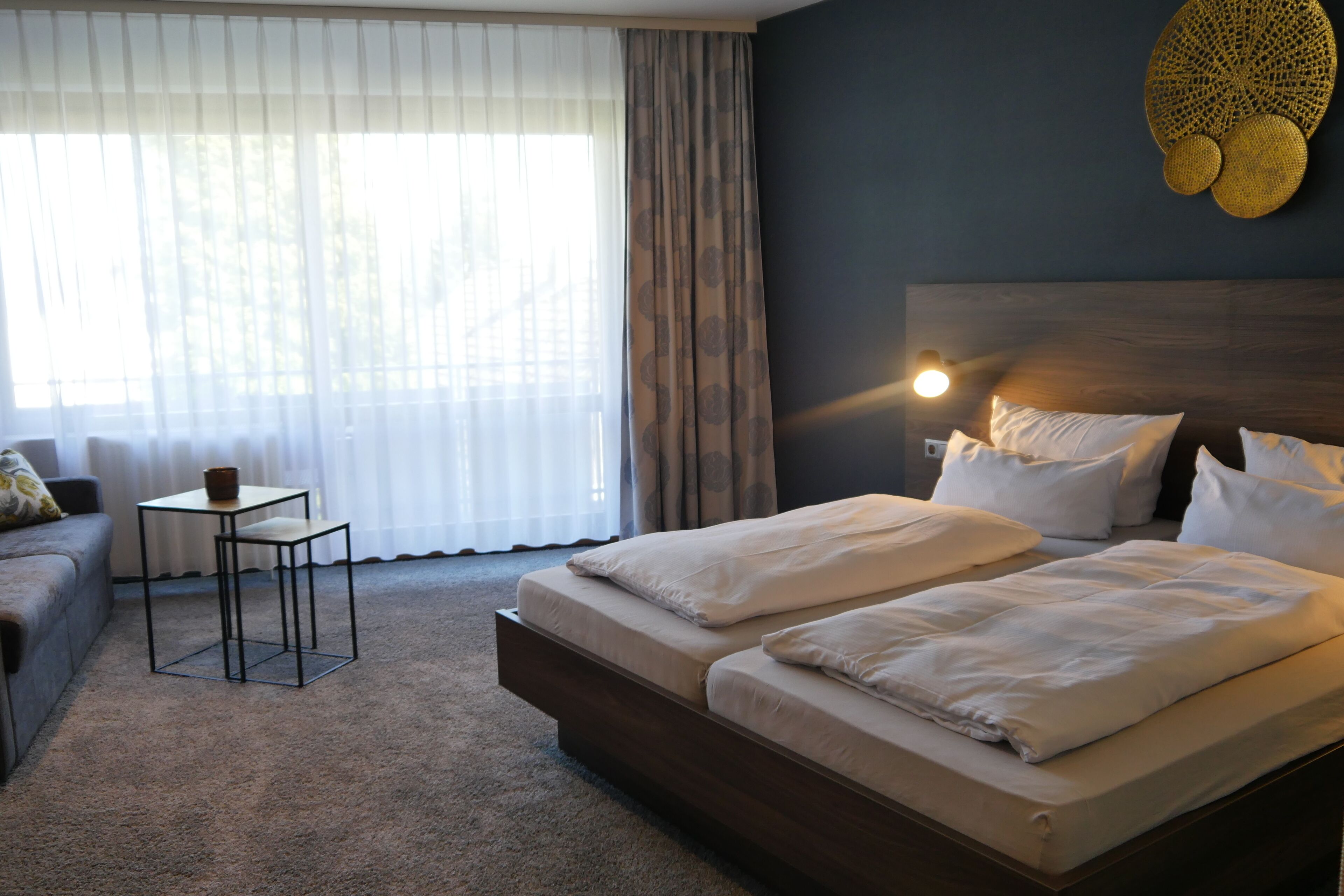 Ein Schlafzimmer mit Bett, kleinem Tisch, Sofa und einem großen Fenster mit Vorhängen.