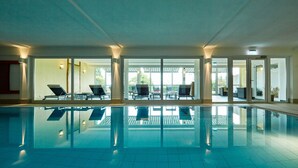 A large indoor swimming pool with clear glass walls, allowing views of the outside. There are lounge chairs and tables near the pool, and a door labeled "Routage" is visible. - NaturKulturHotel Stumpf (Neunkirchen)