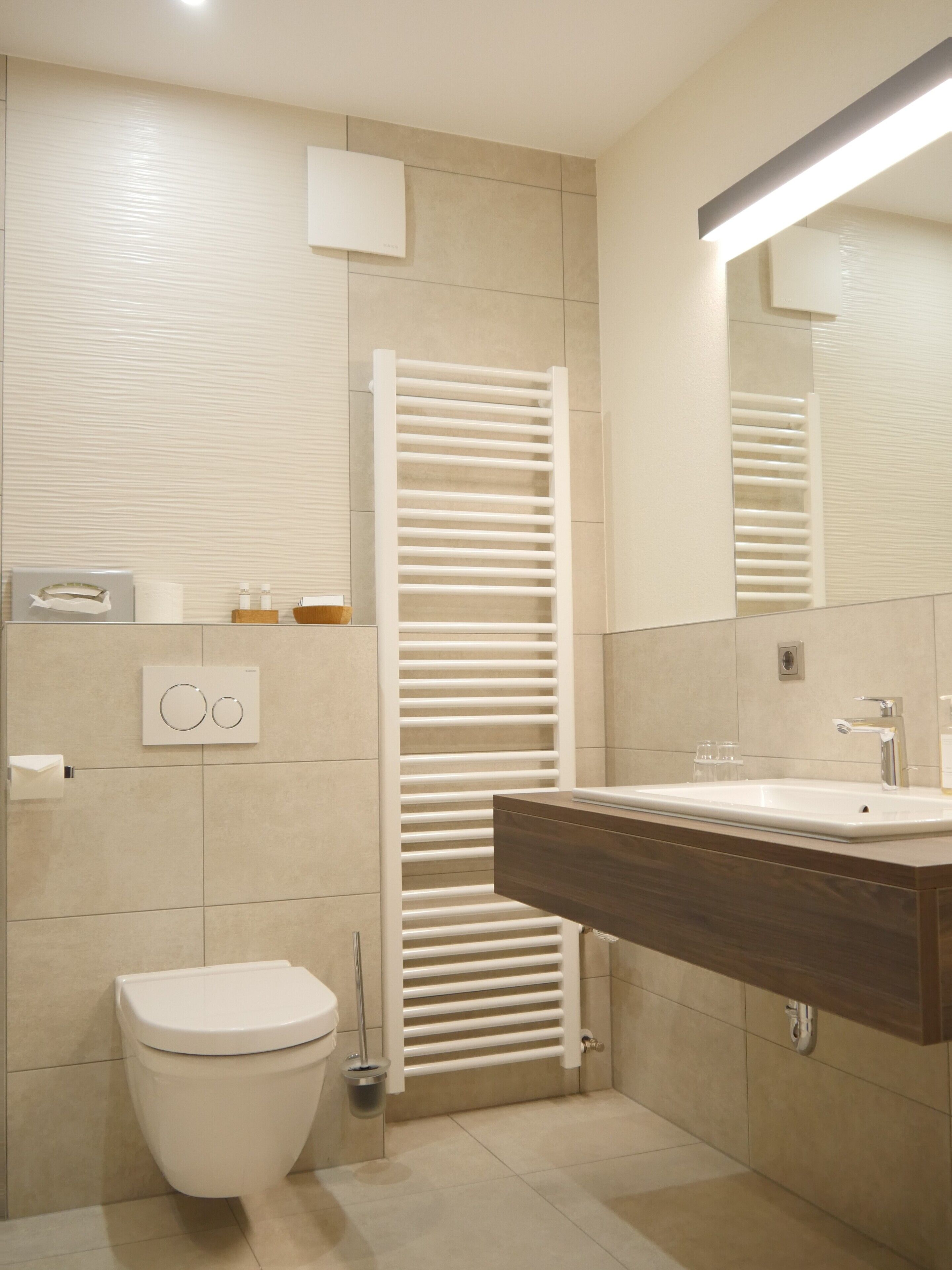 Un baño moderno con inodoro blanco, lavamanos empotrado con encimera de madera, toallero y espejo sobre el lavamanos.