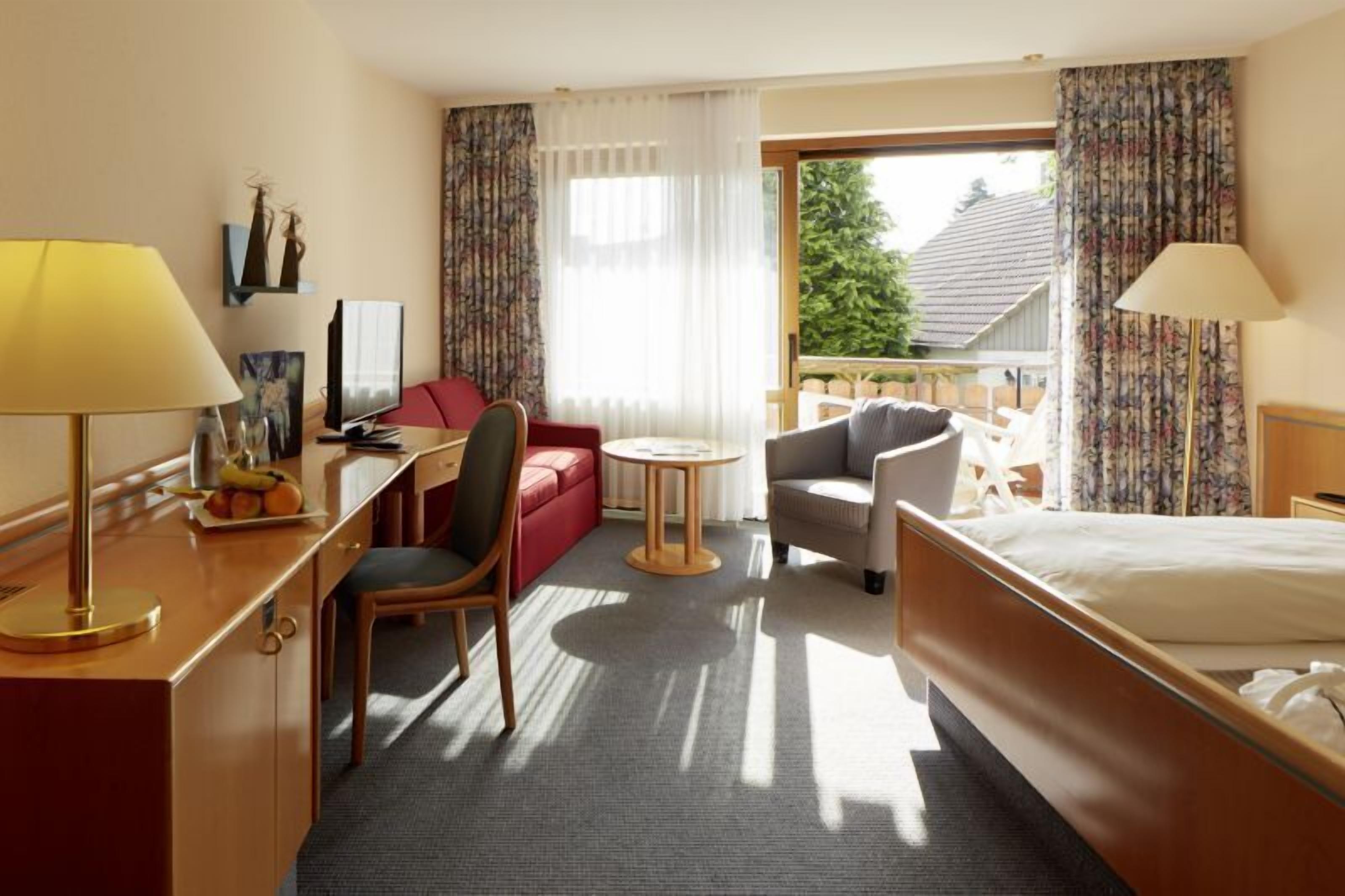 A hotel room with a bed, a desk with a lamp, a red sofa, a round table, and a chair.