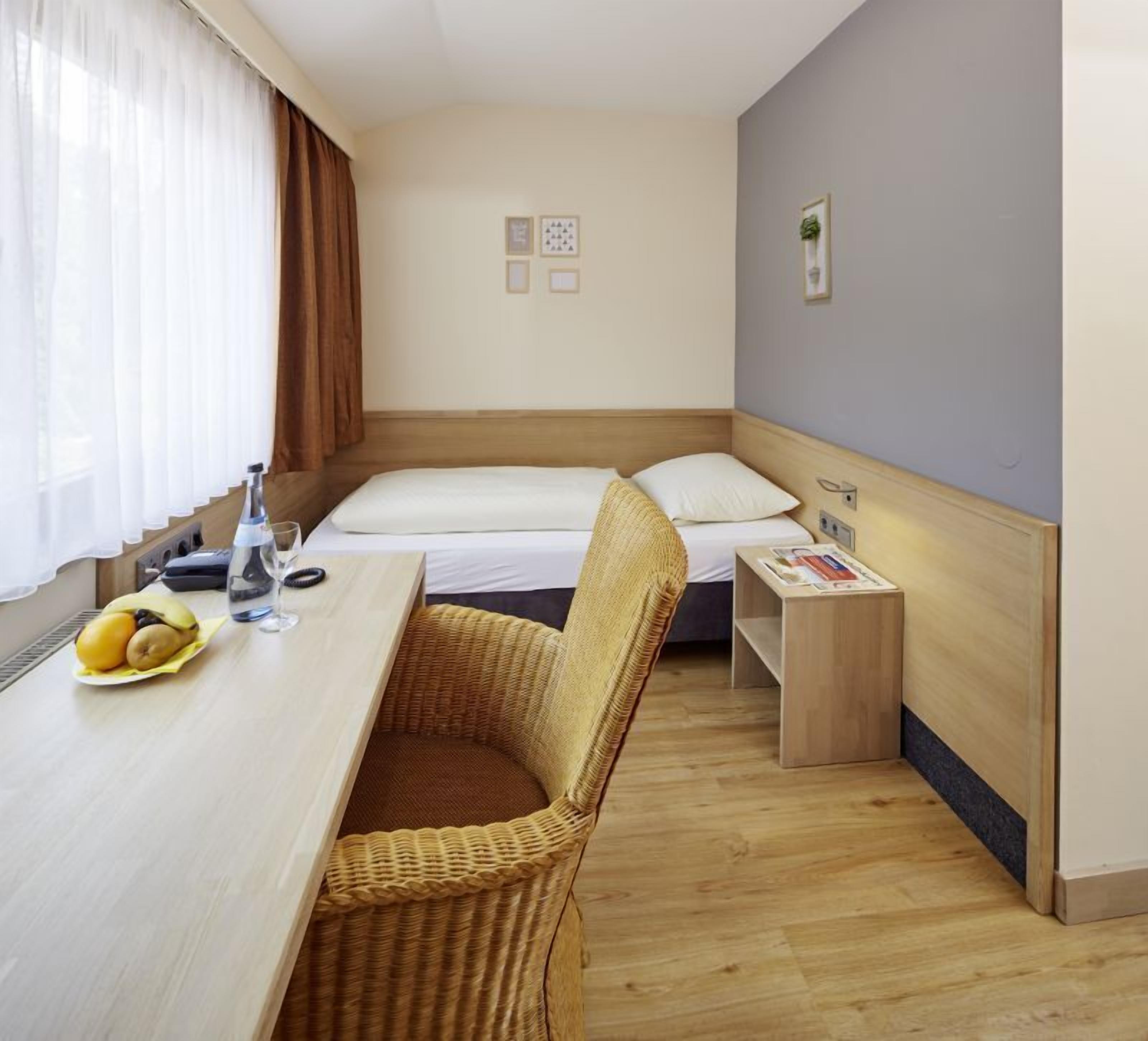 A small hotel room with a single bed, a wooden desk with a chair, a small table with a fruit bowl, and a bedside table with a lamp and a magazine.