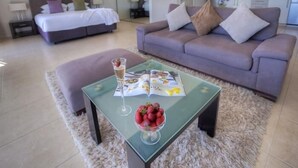 A modern living room with a sofa, a glass coffee table with a magazine and a bowl of strawberries, and a bed in the background.