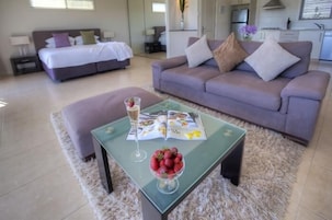 A modern living room with a sofa, a glass coffee table with a magazine and a bowl of strawberries, and a bed in the background.