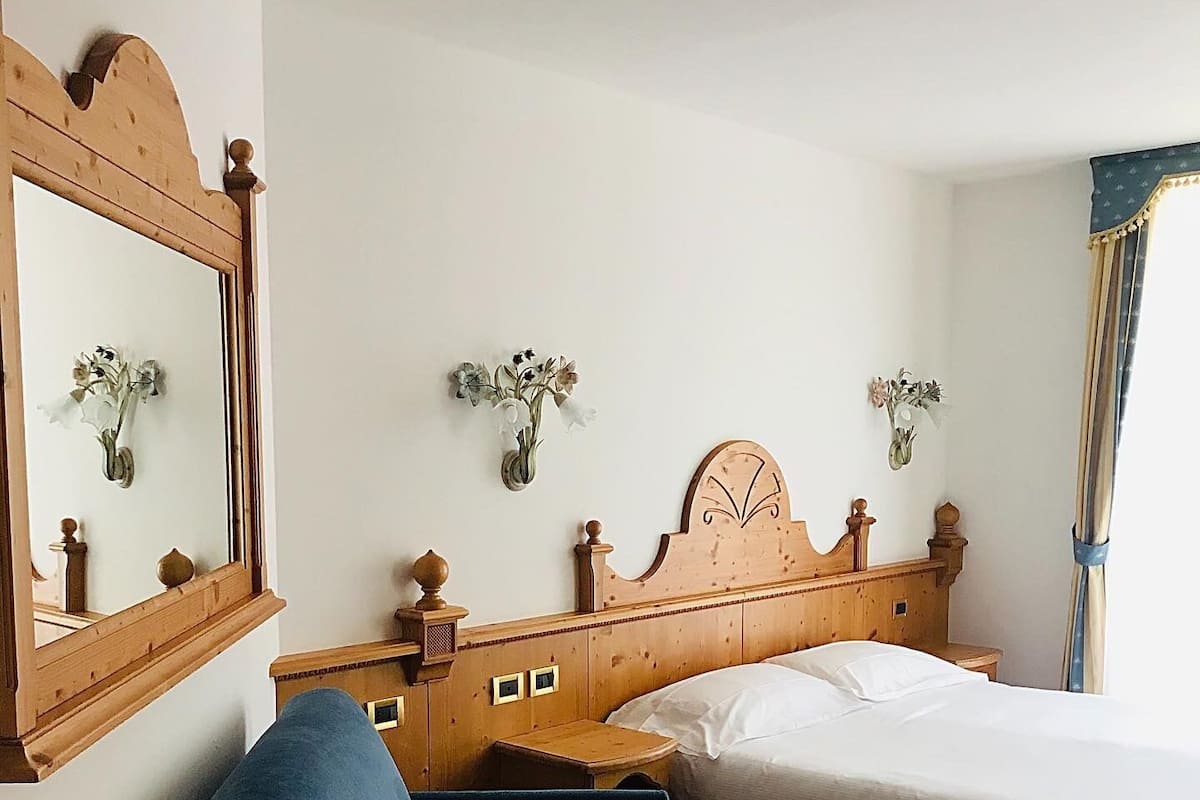 Une chambre à coucher avec une tête de lit en bois, un lit, un fauteuil bleu, une petite table et des compositions florales accrochées au mur.