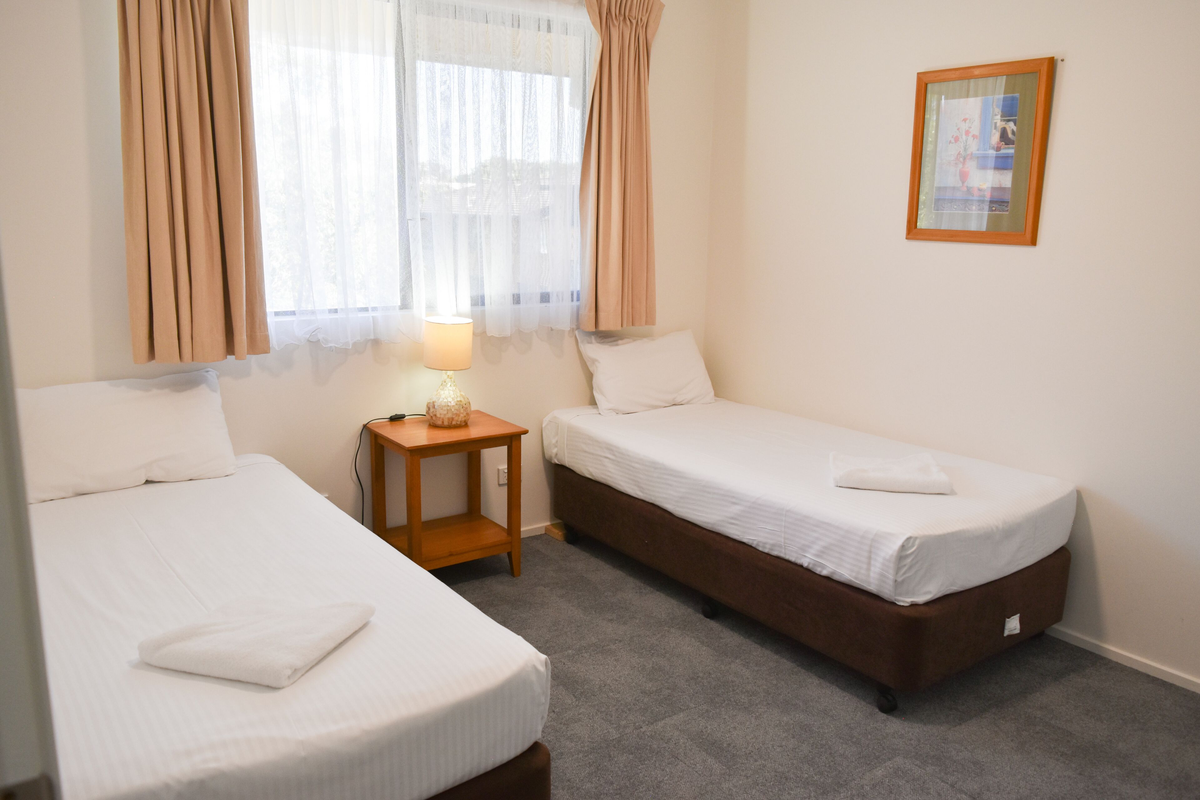 A hotel room with two single beds, a bedside table with a lamp, and a framed picture on the wall.