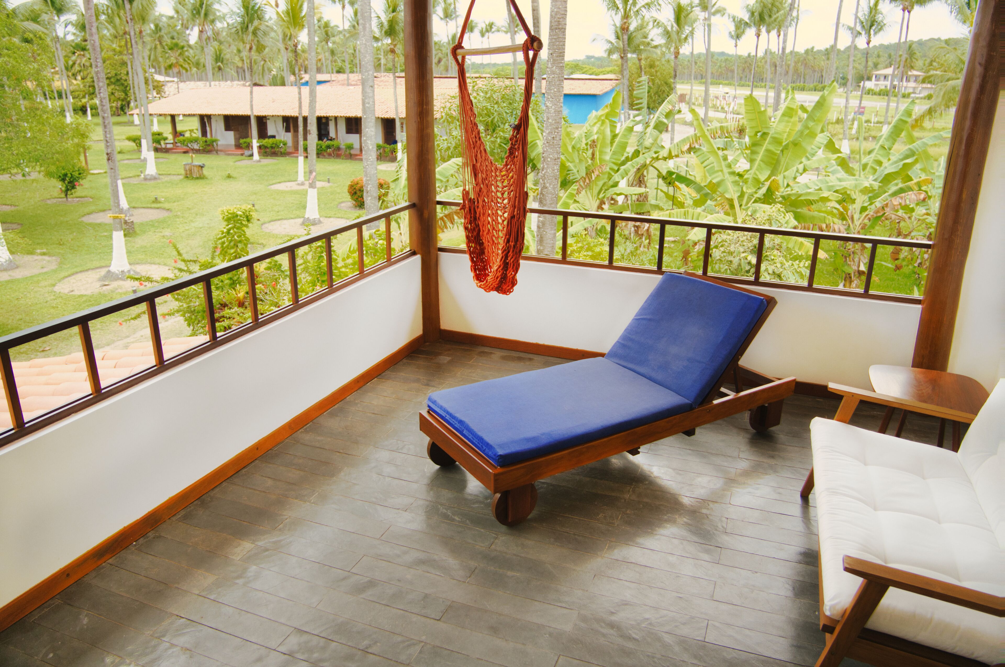 A balcony with a blue cushioned chair, a hanging hammock, and a view of a garden with palm trees.