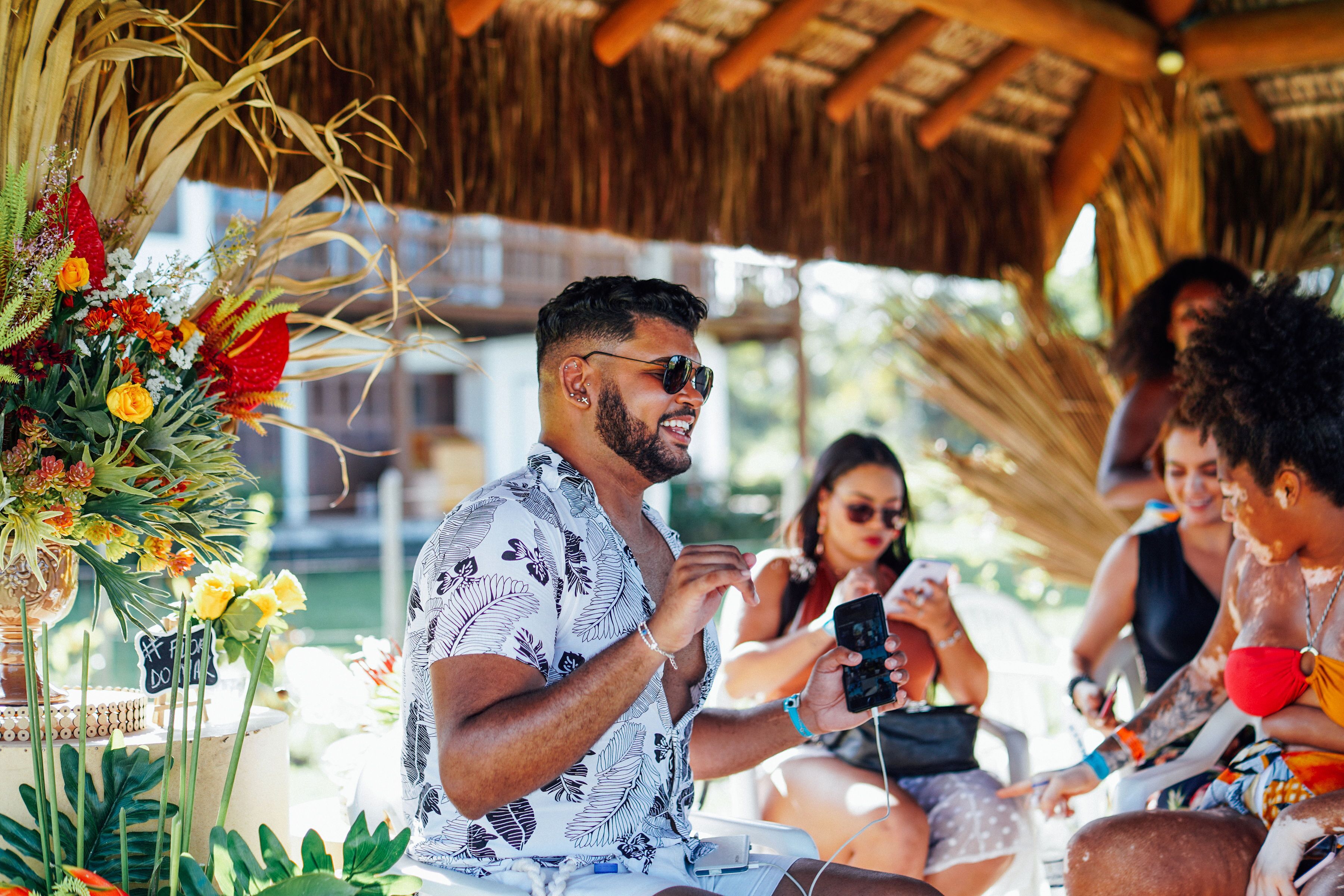 A tropical outdoor setting with a thatched roof structure, a table with a floral arrangement, and people sitting and using mobile devices.