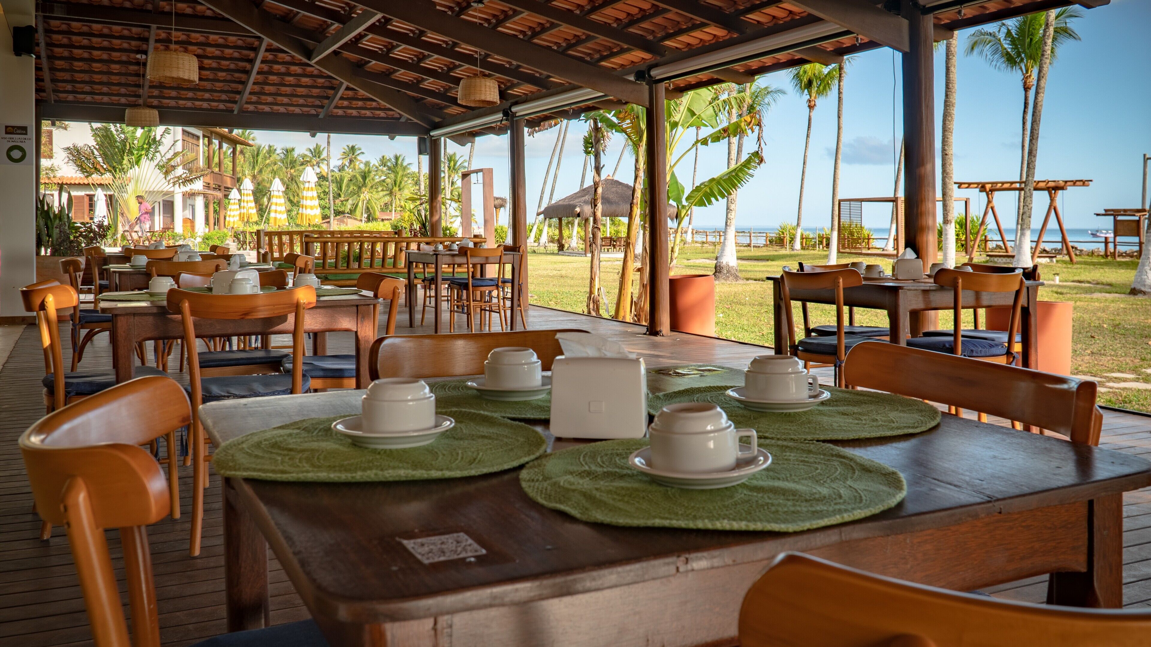 Un área de comedor al aire libre con mesas preparadas para el servicio, rodeada de palmeras y vistas al océano.