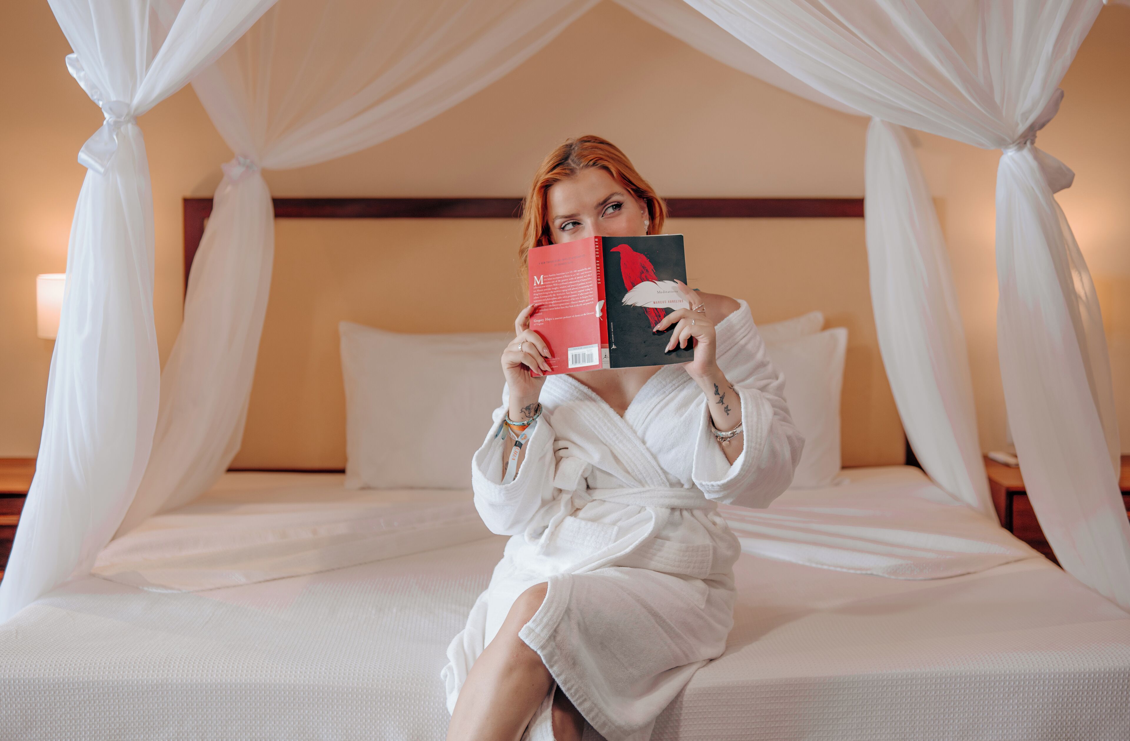 A person sitting on a bed with a canopy, holding a book.