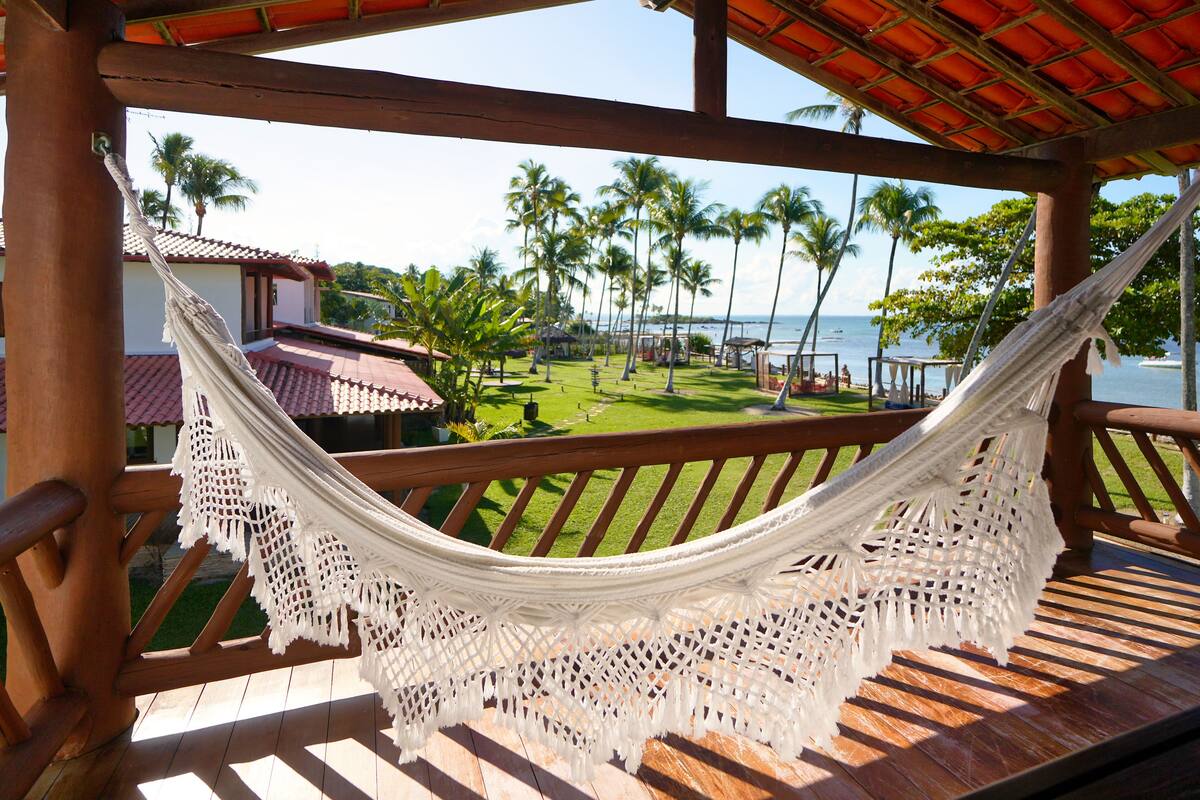 A hammock on a wooden deck with a view of a beach and palm trees.