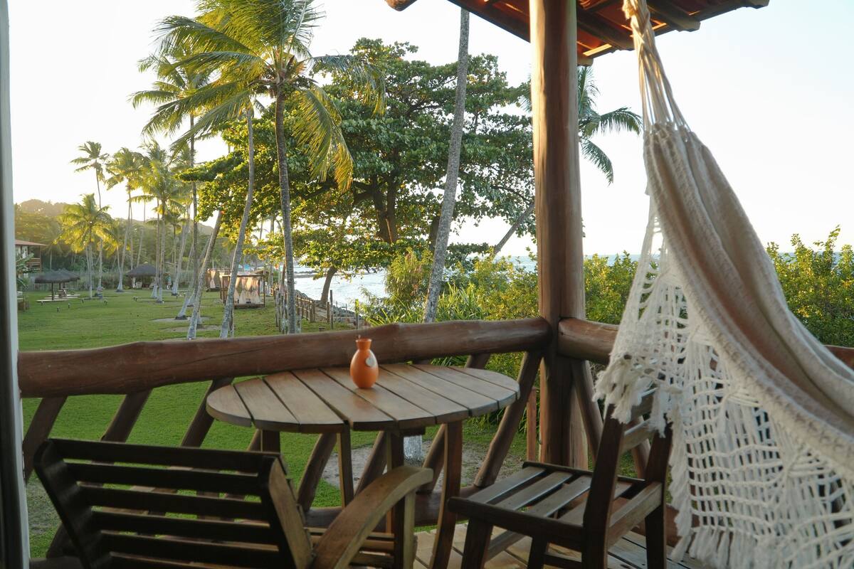 A wooden deck with a hammock, a table with a vase, and a view of palm trees and a beach.