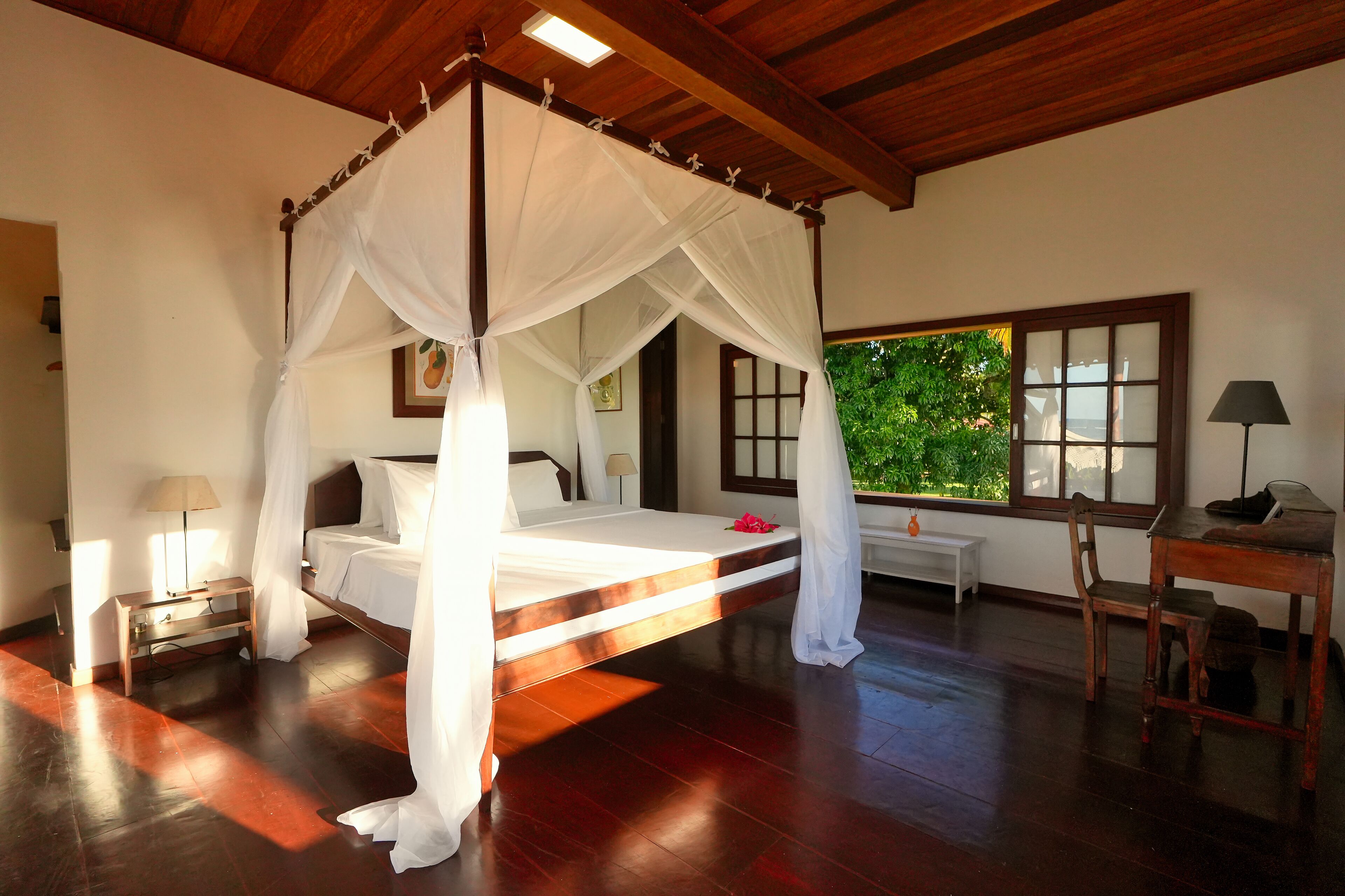 A bedroom with a four-poster bed, a wooden desk, a nightstand, and a window with a view of greenery.