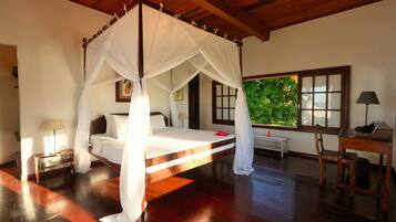 A bedroom with a four-poster bed, a wooden desk, a nightstand, and a window with a view of greenery.