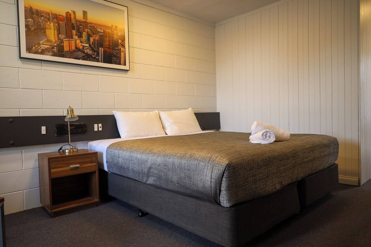 Habitación de hotel impecable con una cama grande, una mesita de noche con lámpara y un cuadro de paisaje urbano en la pared.