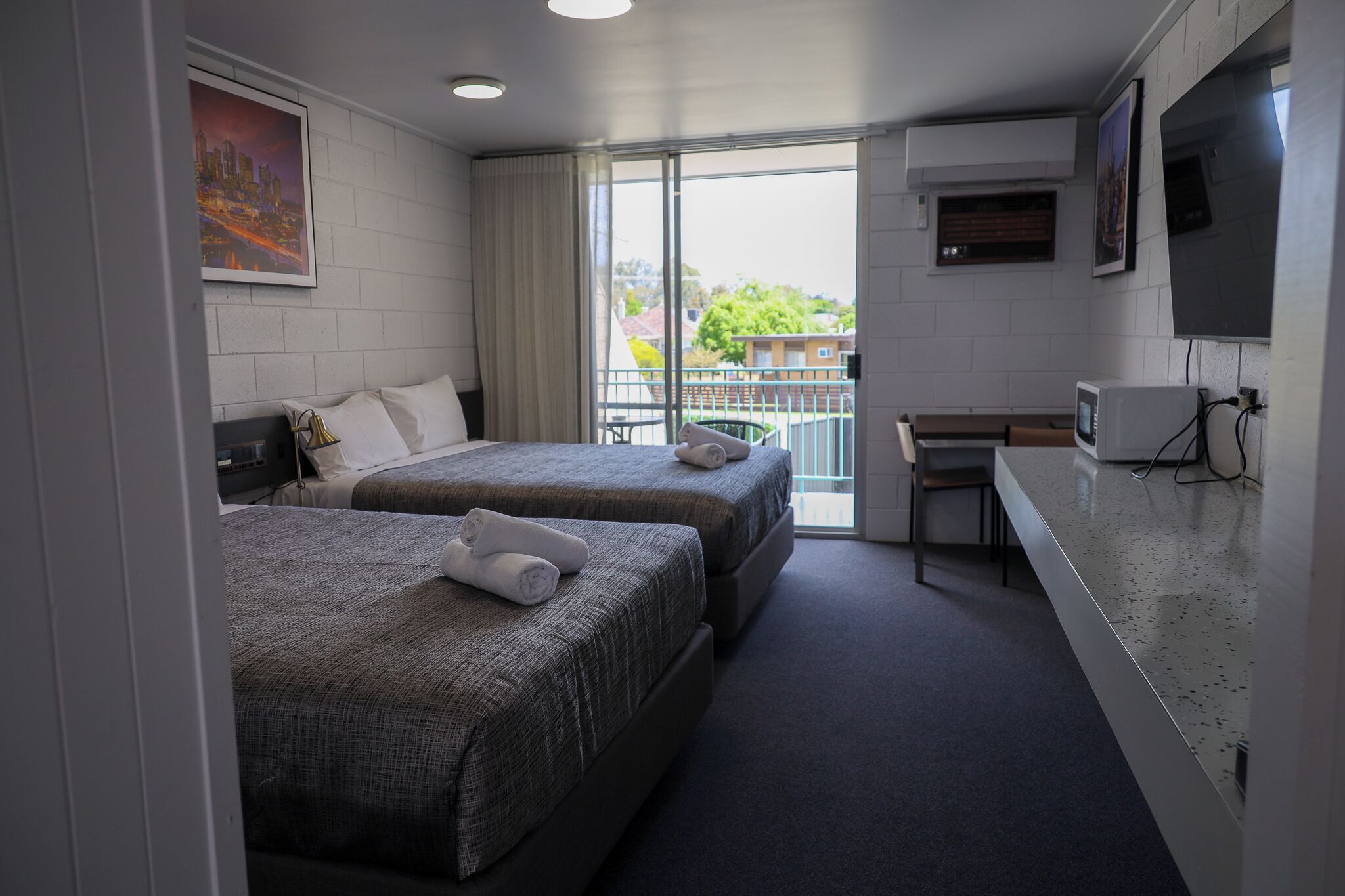 A hotel room with two beds, a desk, a TV, and a balcony with a view of buildings and trees.