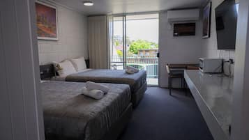 A hotel room with two beds, a desk, a TV, and a balcony with a view of buildings and trees.