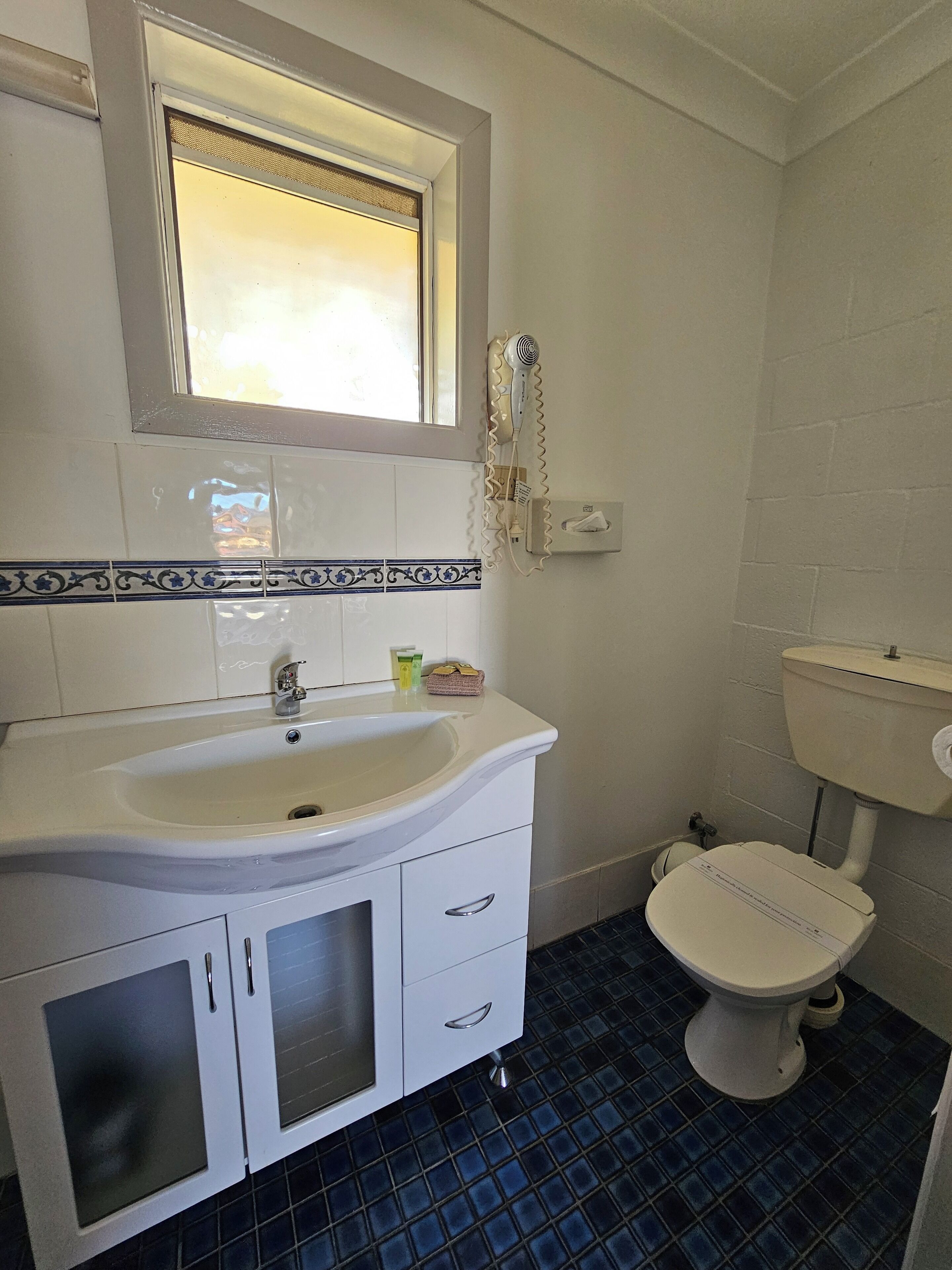 A bathroom with a white sink, a mirror, a window, a toilet, and blue-tiled flooring.