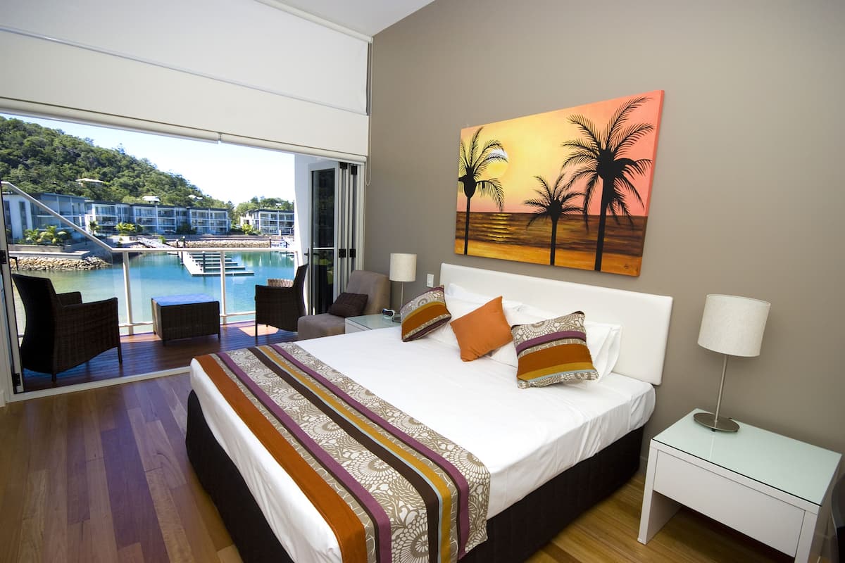 A hotel room with a large bed, a painting of palm trees, a view of a pool, and a balcony with wicker chairs.
