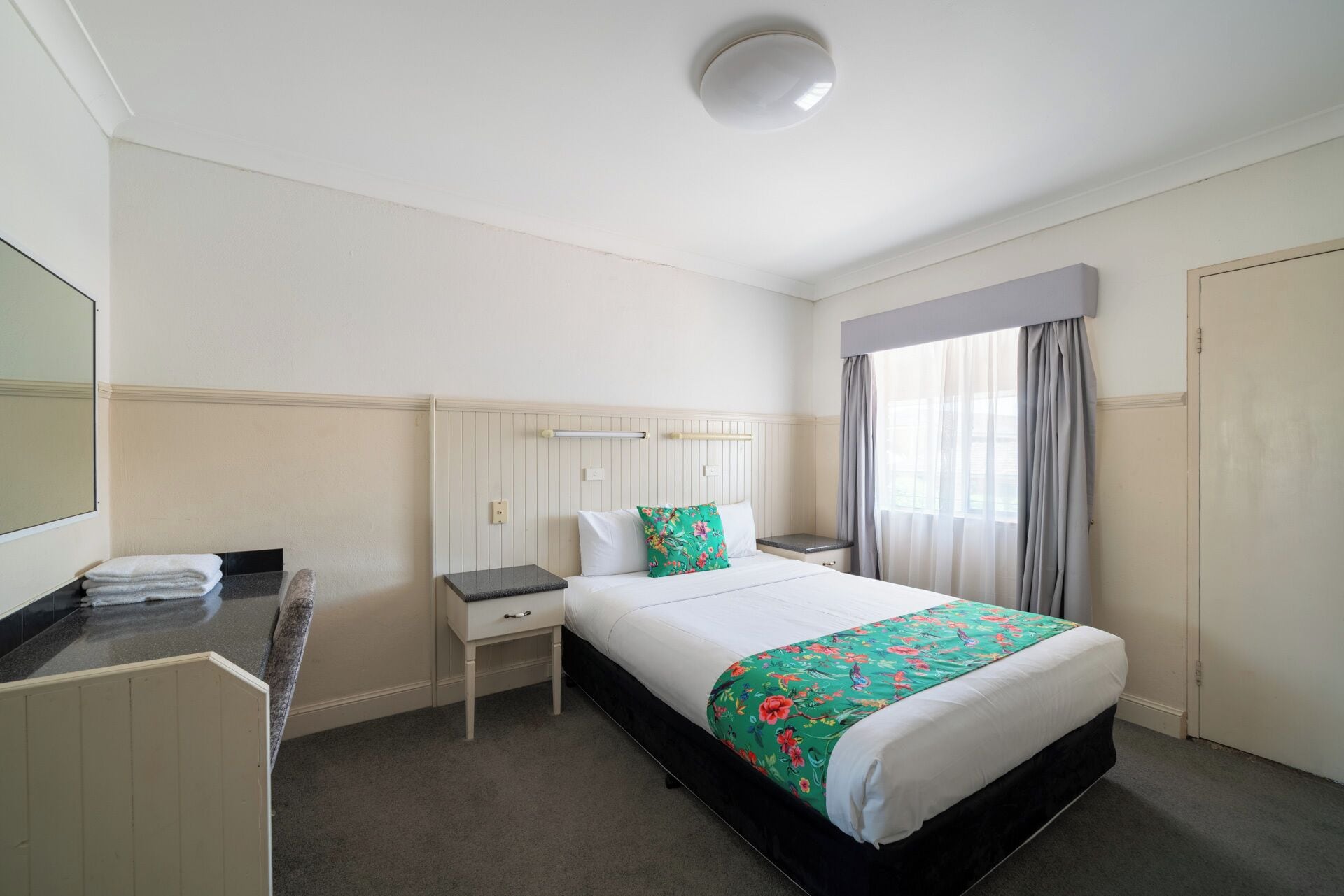 Habitación de hotel con cama, mesita de noche, escritorio con silla, espejo y ventana con cortinas.