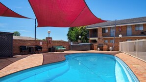 Piscina con una vela de sombra roja, mobiliario exterior y un edificio de ladrillo al fondo.