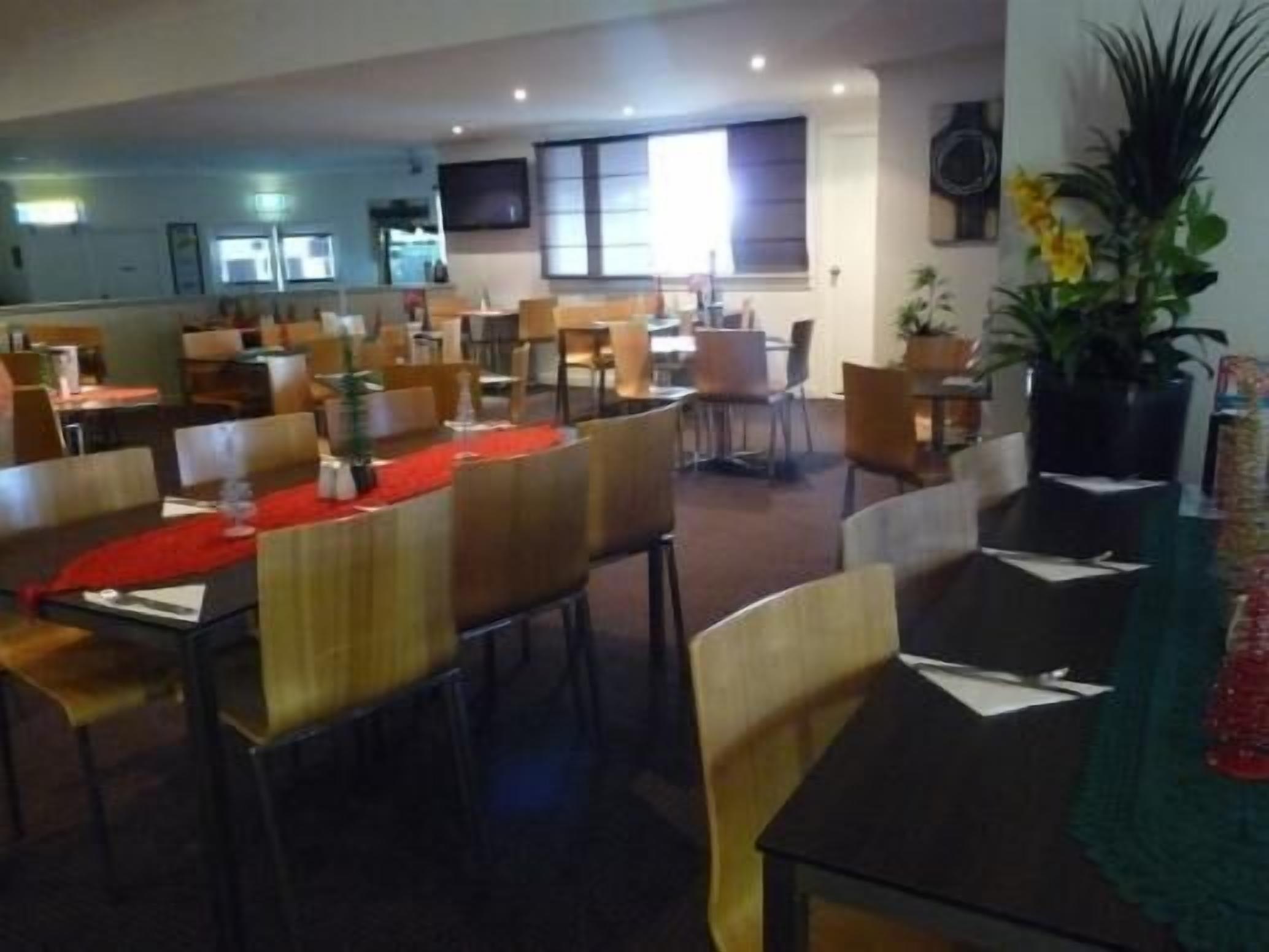 A spacious dining area with multiple tables and chairs, a kitchen in the background, and a large potted plant.