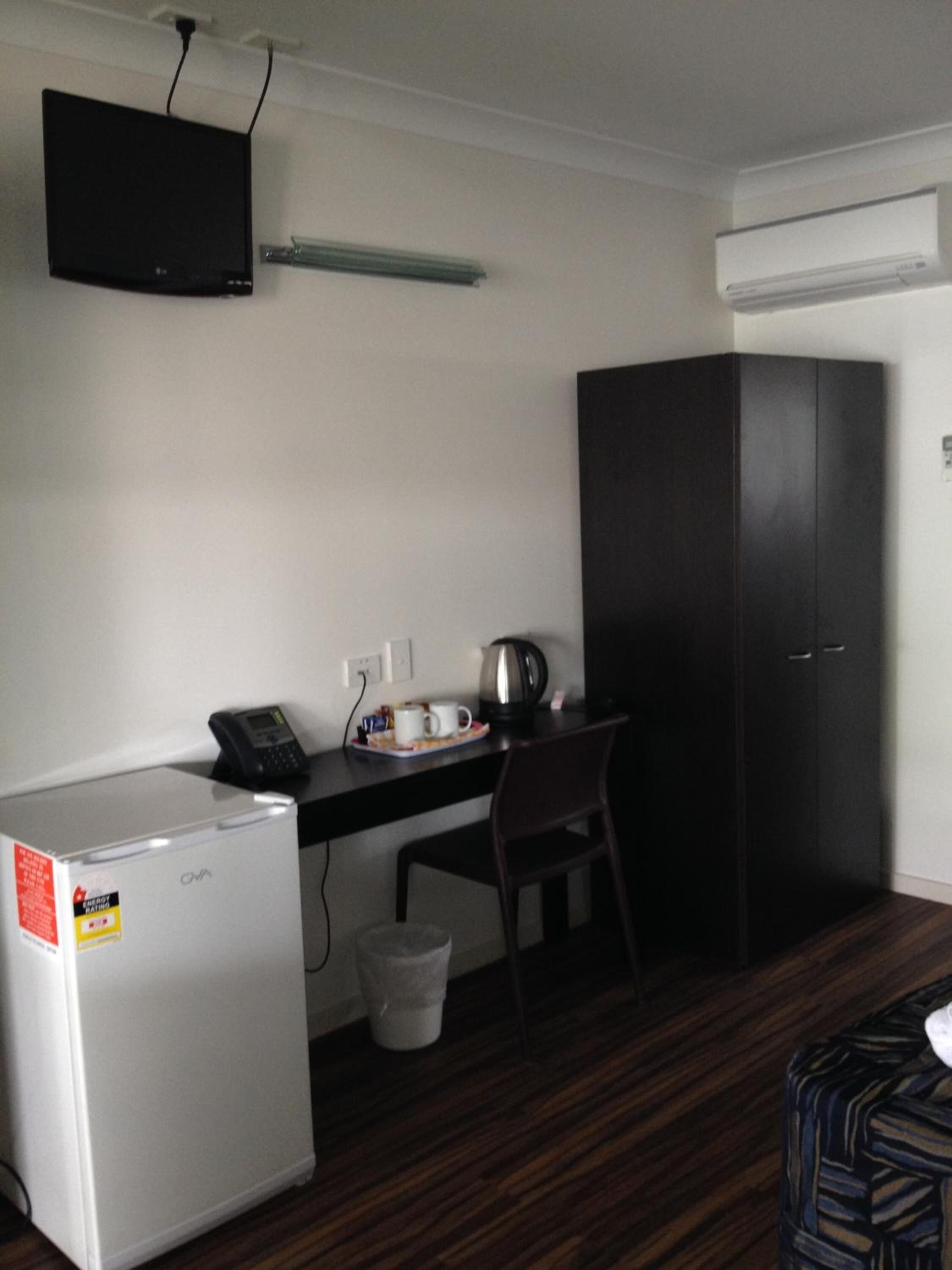 A compact hotel room with a kitchenette, a desk with a chair, a dark wardrobe, and a small trash bin.