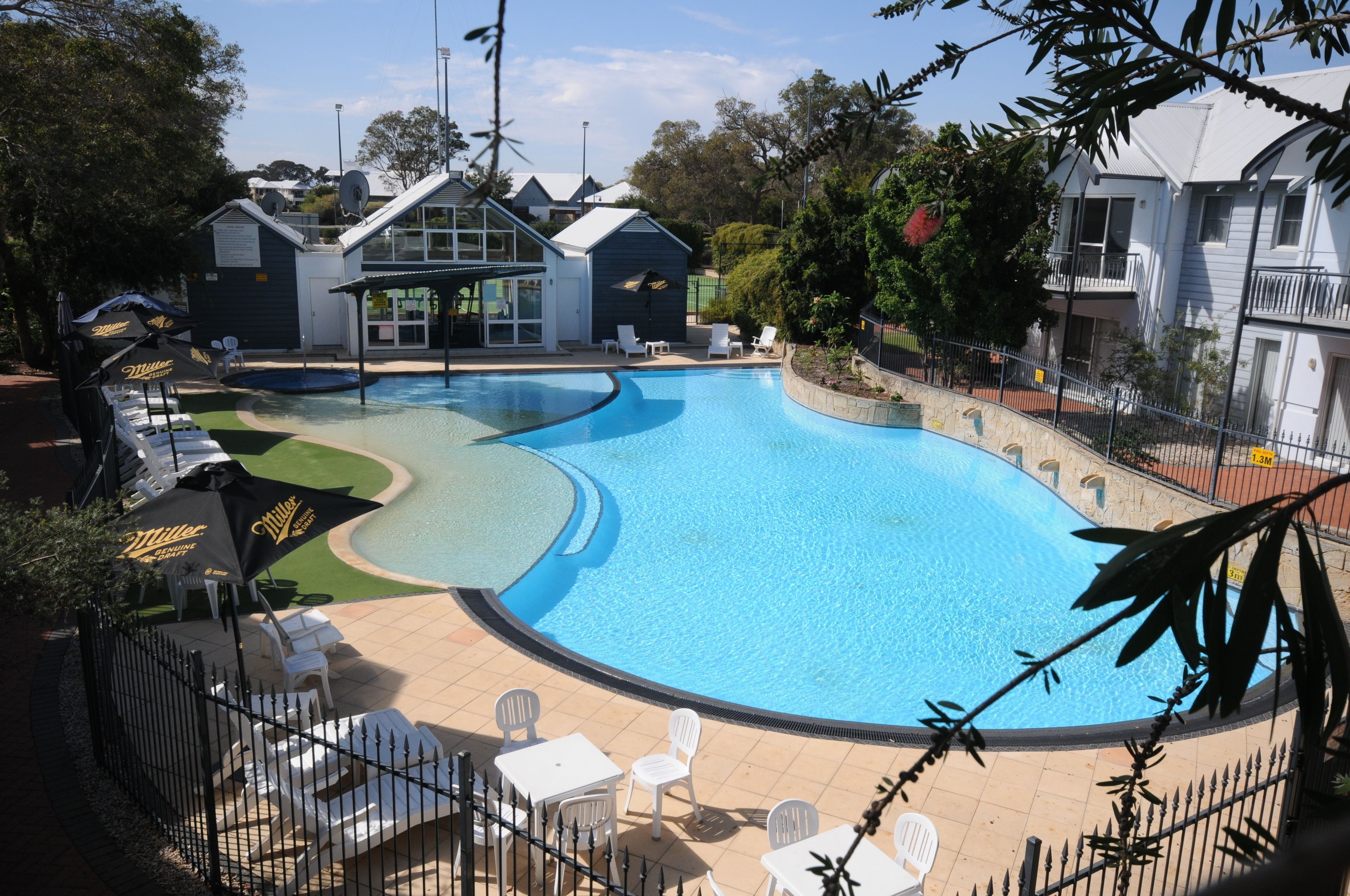 Piscina con sillas de descanso y sombrillas, rodeada por una valla y casas.