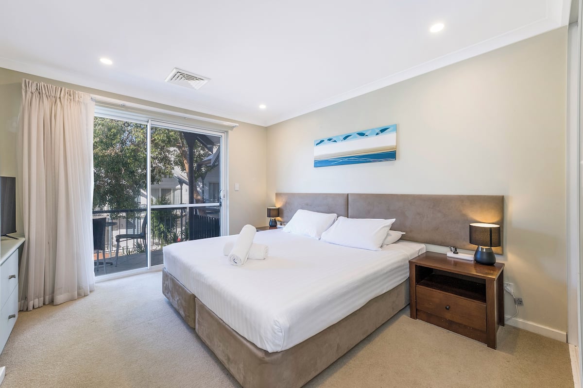 A bedroom with a large bed, a nightstand, a TV, and a sliding glass door to a balcony.