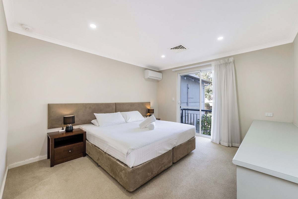 A bedroom with a large bed, a nightstand, a desk, and a balcony with curtains.