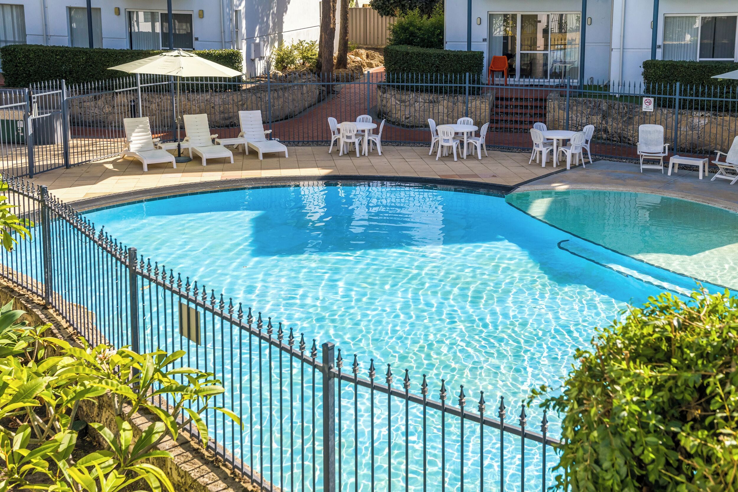 A swimming pool with a metal fence, surrounded by white lounge chairs and tables.