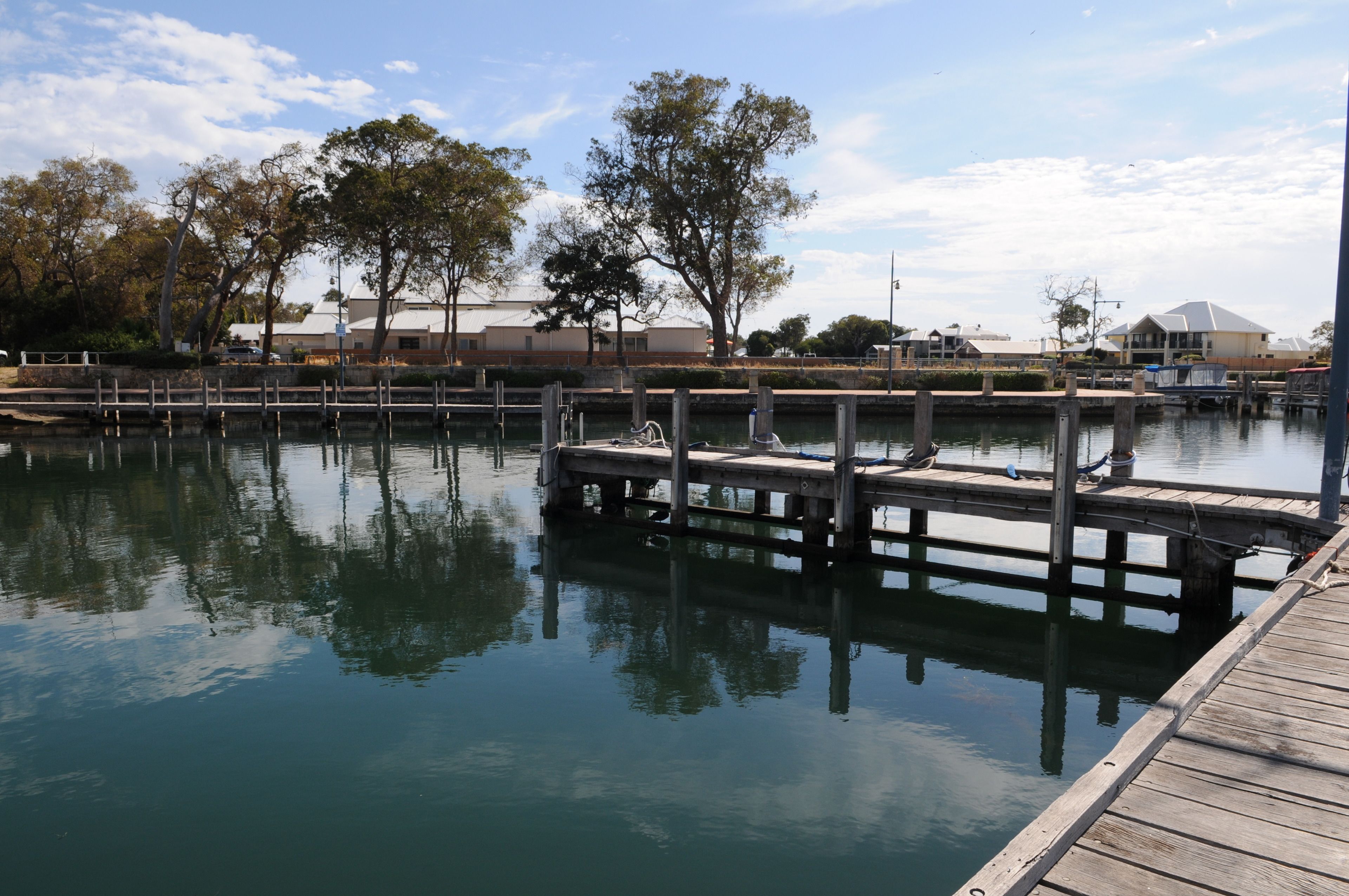 A calm waterfront with a wooden dock extending into the water, surrounded by trees and buildings in the background.