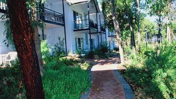 A pathway leading to a white building with balconies, surrounded by greenery and trees.