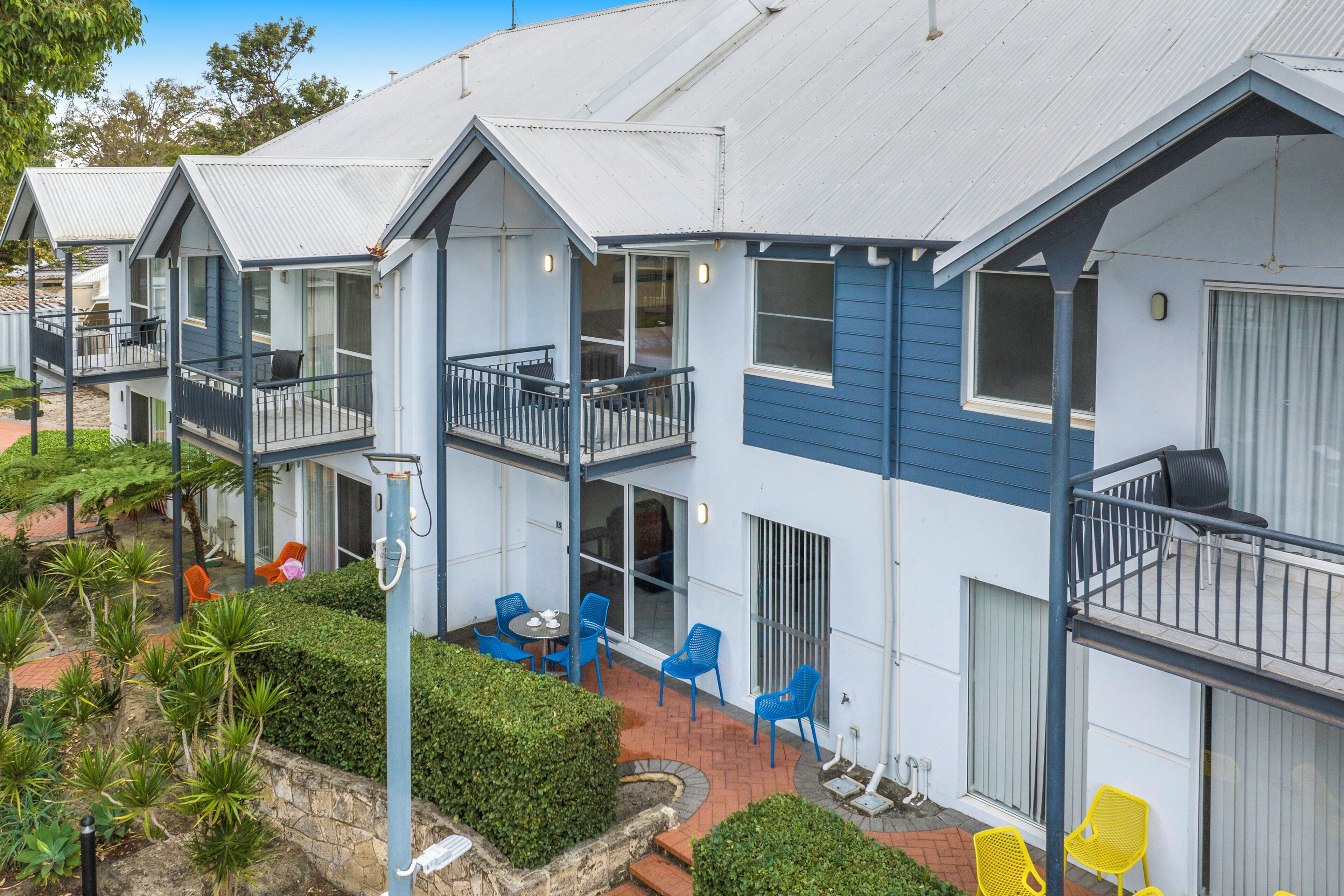 A row of blue and white buildings with balconies, outdoor seating, and a landscaped garden area.