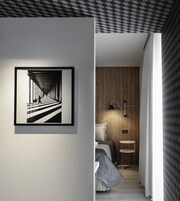Una habitación de hotel moderna con una fotografía en blanco y negro enmarcada en la pared, un armario de madera, una mesita de noche con una lámpara y una cama con una manta gris.