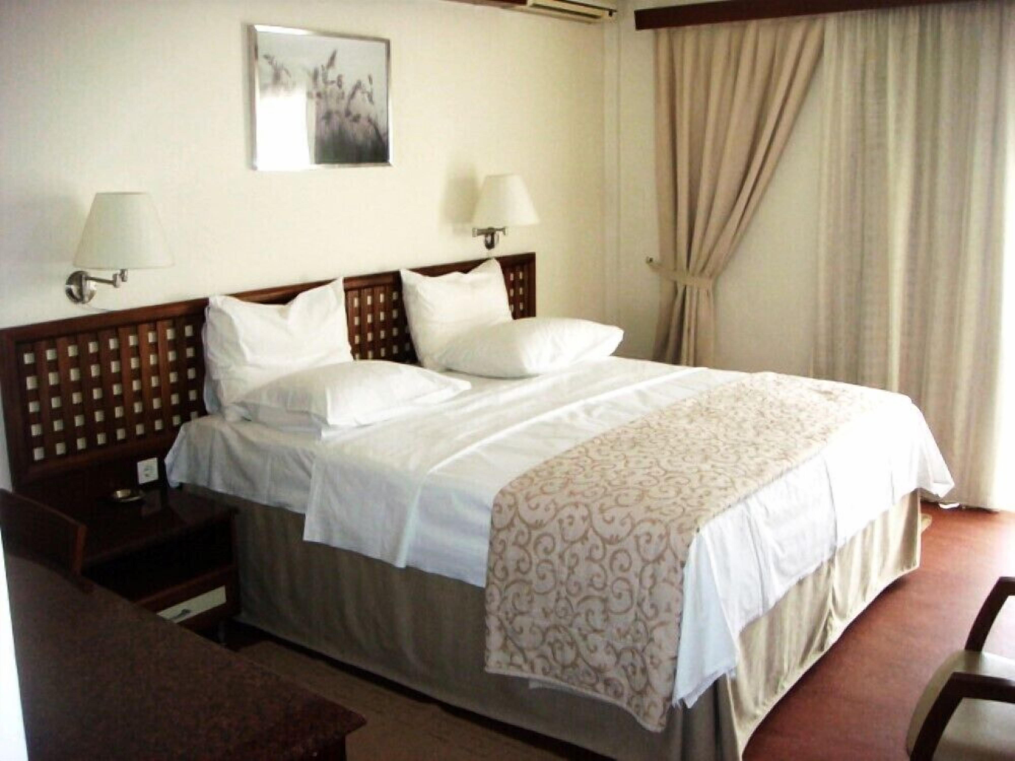 A hotel room with a large bed, two bedside tables, a nightstand, a desk, a chair, a lamp, a wall-mounted picture, and a window with curtains.