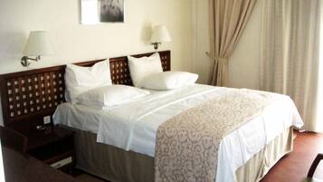 A hotel room with a large bed, two bedside tables, a nightstand, a desk, a chair, a lamp, a wall-mounted picture, and a window with curtains.