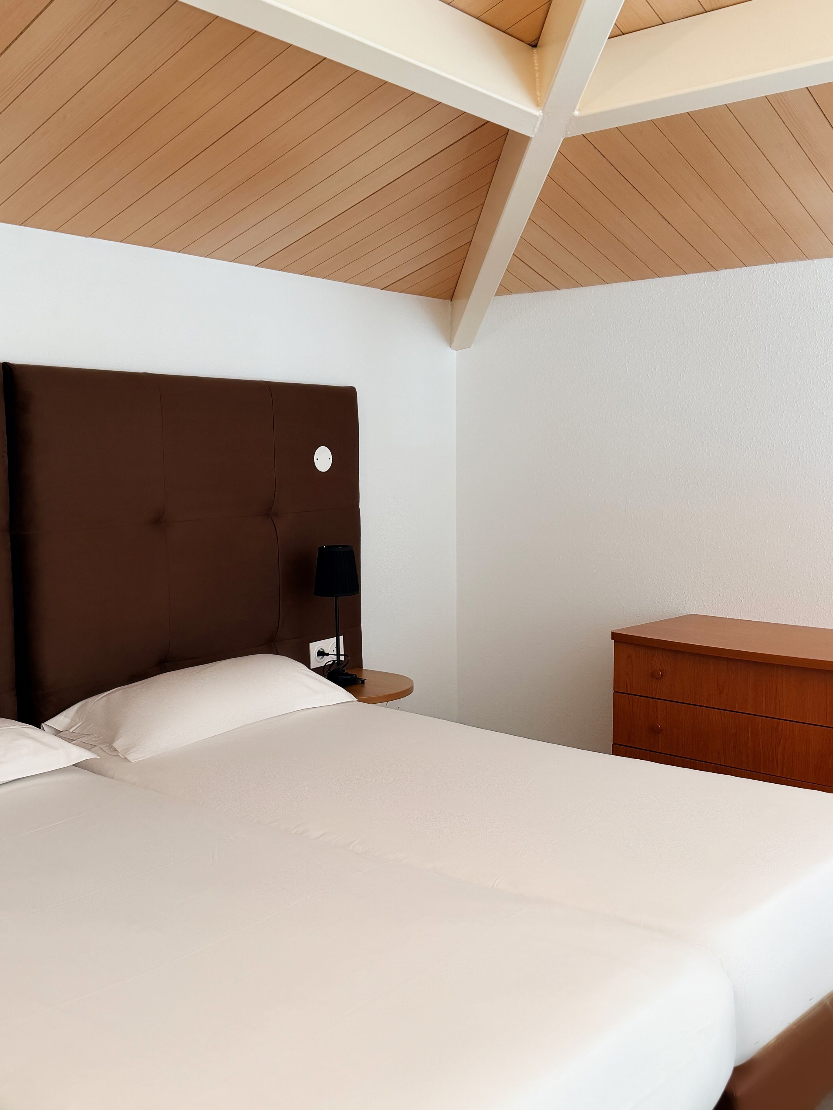 Una habitación de hotel con una cama, una mesita de noche, un aparador y techo de madera.