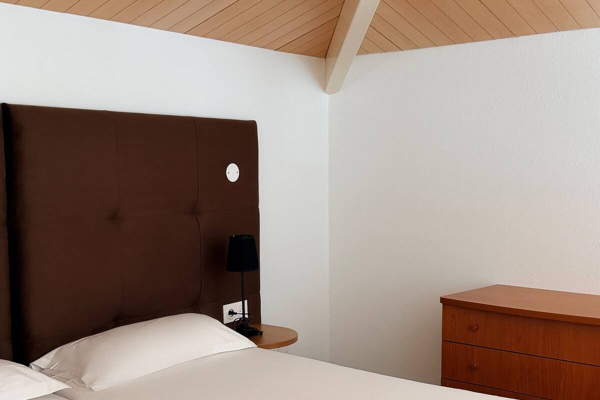 Una habitación de hotel con una cama, una mesita de noche, un aparador y techo de madera.