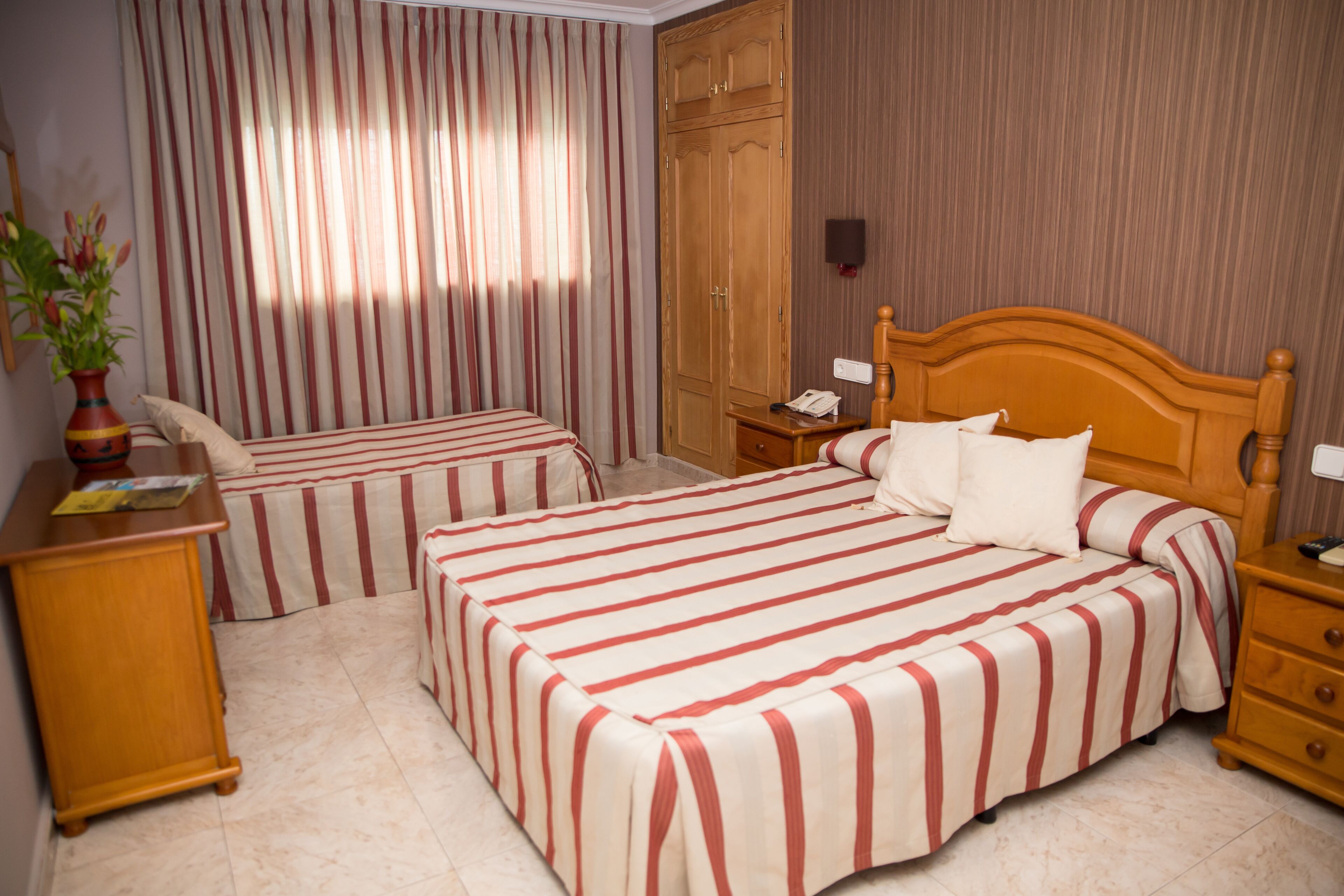 A hotel room with two beds, a wooden wardrobe, a bedside table with a lamp, and a potted plant.