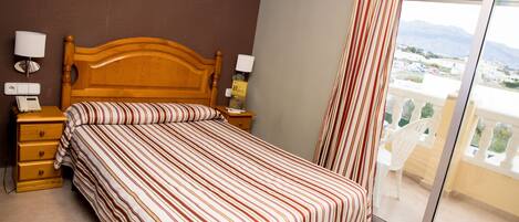 A hotel room with a wooden bed, two bedside tables with lamps, a striped bedspread, and a view of a balcony with chairs.