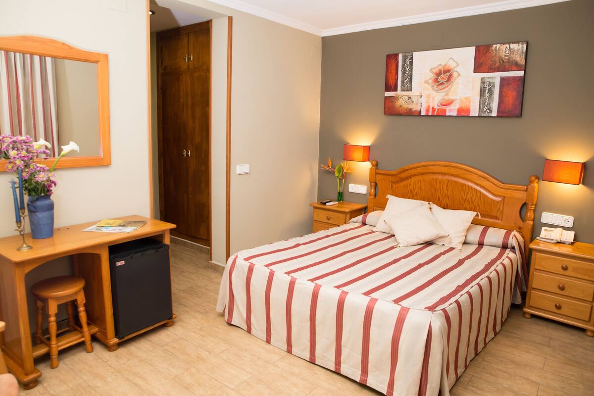 Une chambre d’hôtel équipée d’un lit, d’un bureau, d’une chaise, d’un petit réfrigérateur et d’une table de chevet avec une lampe.