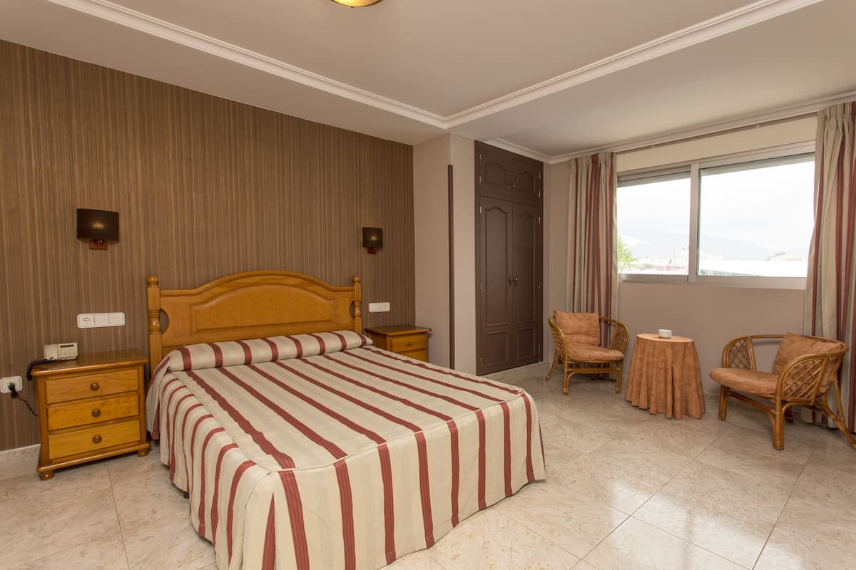 Une chambre d’hôtel avec un grand lit, deux tables de chevet, une armoire en bois, deux fauteuils en osier, une petite table ronde et une fenêtre avec des rideaux.