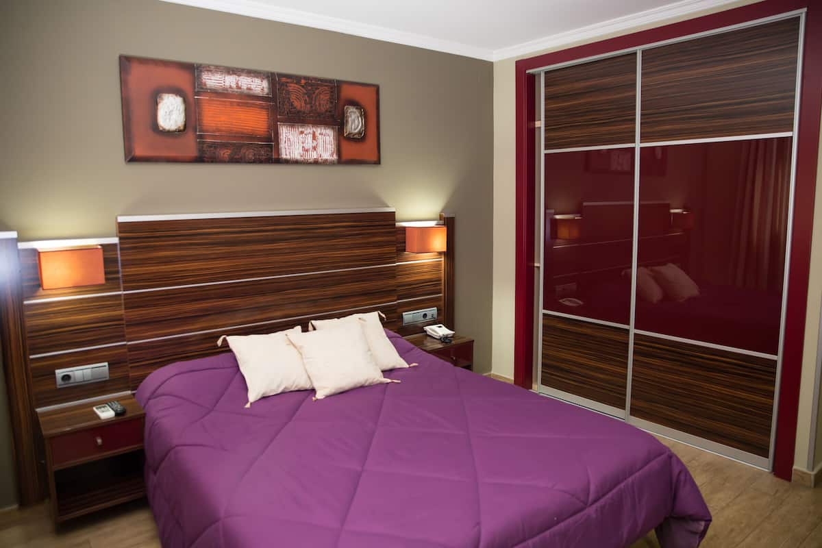 Une chambre à coucher avec une tête de lit en bois, une couvre-lit violette, deux tables de chevet avec des lampes et un grand miroir.