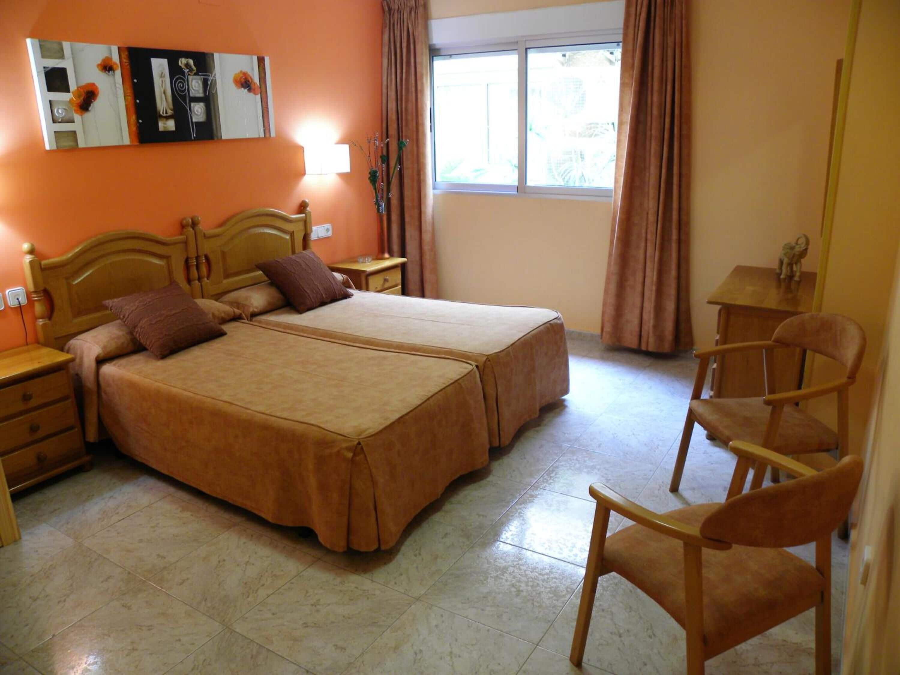A double bed with a wooden headboard, a nightstand, a chair, and a window with curtains.