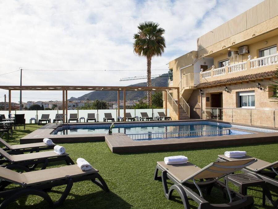 A pool area with lounge chairs and a pergola, surrounded by a building with balconies and a mountain view.