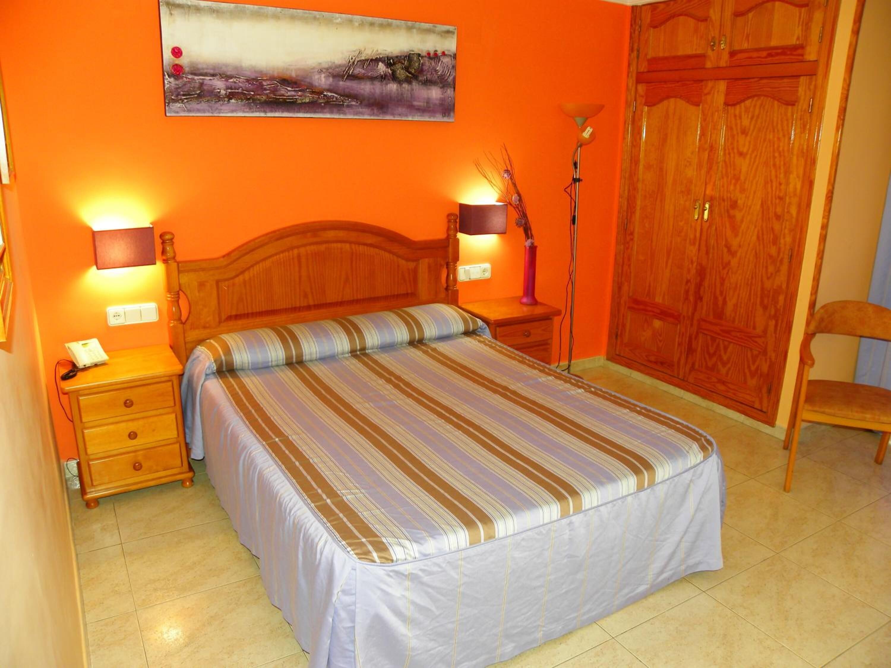 A bedroom with a bed, two bedside tables, a nightstand, a lamp, a chair, and a wardrobe.