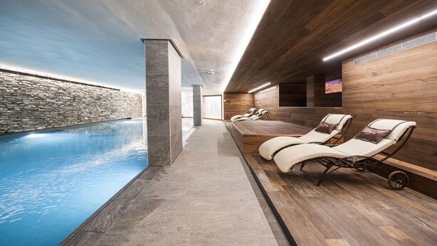 A modern indoor pool area with a stone wall, wooden deck, and lounge chairs. - Hotel Touring Livigno (Livigno)